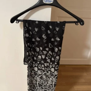 Svart halsduk med grå hjärtan - Säljer en svart halsduk med grå hjärtmönster. Halsduken är tunn och lätt, perfekt som accessoar för att lyfta din outfit. Mönstret består av små och stora hjärtan över hela tyget.