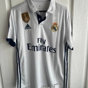 Real Madrid fotbollströja Ronaldo 7 - Säljer en vit Real Madrid fotbollströja med Ronaldo 7 på ryggen. Tröjan har mörkblå detaljer, klubbmärke och Adidas-logga på bröstet samt Fly Emirates-tryck. Kortärmad modell med FIFA-märke och Champions League-märke på ärmen.