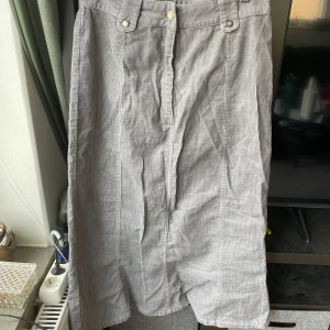 Grå manchesterkjol från Fashion Quality - Säljer en grå manchesterkjol från Fashion Quality med diskreta ränder och breda hällor för bälte. Kjolen har knäppning fram och är i en längre modell som passar perfekt till vardagslooken.