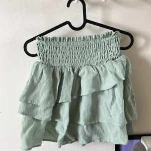 grön kjol - Supersöt ljusgrön kjol från H&M med smock i midjan och flera lager volanger. Kjolen är kort och har en luftig känsla, perfekt för varmare dagar. Enkel att matcha med olika toppar för en trendig look. Nästan aldrig använd