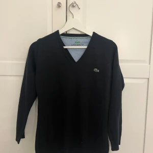 Svart v-ringad tröja från Lacoste - Säljer en klassisk svart v-ringad tröja från Lacoste med den ikoniska krokodilloggan på bröstet. Tröjan har långa ärmar och är tillverkad i ett mjukt material som passar perfekt till många olika outfits.