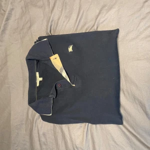 Mörkblå pikétröja från Burberry - Säljer en klassisk mörkblå pikétröja från Burberry med lång ärm och broderad logga på bröstet. Tröjan har en typisk pikékrage och knäppning framtill samt detaljer i beige på insidan av kragen.