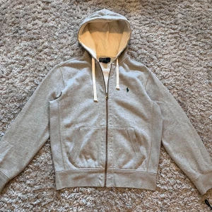 Grå hoodie från Polo Ralph Lauren - Säljer en klassisk grå hoodie från Polo Ralph Lauren med dragkedja och huva. Tröjan har två fickor framtill och den ikoniska gröna loggan broderad på bröstet. Perfekt för en avslappnad stil.  Det är en liten fläck man ser på sista bilden.