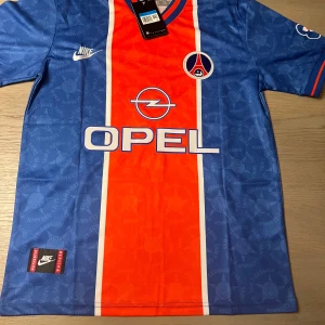 PSG fotbollströja Ronaldinho 10 - Klassisk Paris Saint-Germain fotbollströja från Nike med Ronaldinho och nummer 10 på ryggen. Blå bas med bred röd rand i mitten, vita detaljer och OPEL sponsortryck. Kortärmad modell med klubbmärke och Nike-logga på bröstet.