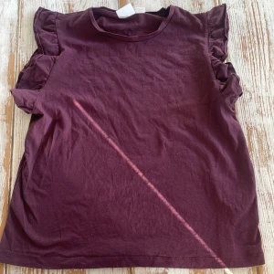 Vinröd topp med volangärmar från Gina Tricot - Säljer en vinröd topp från Gina Tricot med volangdetaljer på axlarna. T-shirten har rund halsringning och är tillverkad i mjuk bomull. Perfekt för dig som vill ha en enkel men söt topp med det lilla extra.