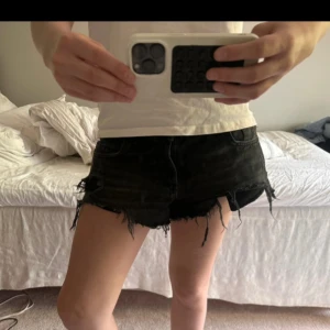 Svarta jeansshorts från Zara - Snygga svarta jeansshorts från Zara med låg midja och råa, slitna kanter. Shortsen har flera coola hål och fransar för en edgy vibe. Klassisk femficksmodell med knapp och dragkedja. Perfekta till sommaren för en avslappnad och trendig look.
