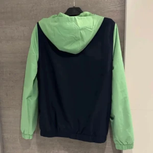 Lacoste vindjacka i blått och grönt - Snygg vindjacka från Lacoste Sport med marinblå kropp och ljusgröna ärmar och huva. Jackan har dragkedja framtill, elastiska muddar och klassisk Lacoste-logga på bröstet. Perfekt för dig som gillar sportig och stilren look.