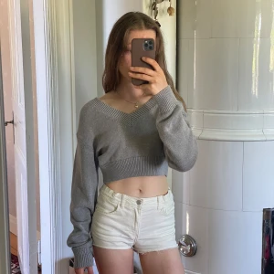 Grå croppad stickad tröja från Gina Tricot -  ljusgrå croppad stickad tröja från Gina Tricot med v-ringning, långa ärmar och bred ribbad kant nedtill och vid ärmsluten. 