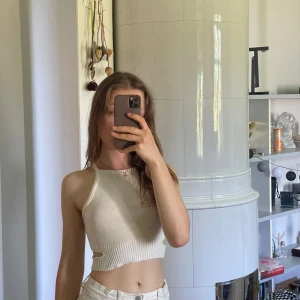 Vit stickad croptop med knyt i ryggen -  vit stickad croptop med korsade band i ryggen som knyts. Toppen är ärmlös och har en tight passform, perfekt för varma dagar.