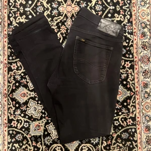 Lee Luke 32/32 svarta - Svarta jeansbyxor från Lee med klassisk femficksdesign och Lee-logga på bakfickan. Byxorna har raka ben och normal passform, tillverkade i ett mjukt denimtyg. Perfekta för en stilren och enkel look.