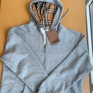 Grå zip-hoodie från Burberry med rutigt foder - Grå hoodie med dragkedja från Burberry. Klassisk huva fodrad med Burberrys ikoniska rutig mönster i beige, svart och rött. Broderad logga på bröstet och justerbar dragsko i huvan. Perfekt för en stilren och avslappnad look.