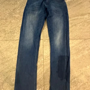 Replay blå jeans straight fit - Snygga blå jeans från Replay med klassisk femficksdesign och diskreta slitningar framtill. Jeansen har raka ben och normal passform, med Replay-logga på fickan och bak på linningen. Perfekta för en avslappnad och stilren look.