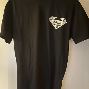Svart t-shirt från Prada med logga  - Svart t-shirt från Prada med ikonisk logga i vitt på bröstet. Klassisk passform och rund halsringning. Tillverkad i mjuk bomull, perfekt för en clean och stilren look. Kommer med äkthetsbevis och originaltaggar.
