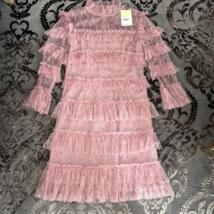 Rosa spetsklänning med volanger från By Malina - Superfin rosa kortklänning i spets från By Malina med volanger över hela klänningen och långa ärmar. Klänningen har hög hals och transparenta partier, vilket ger en romantisk och trendig look. Perfekt för dig som vill sticka ut med en söt och feminin stil. Nypris:3000kr