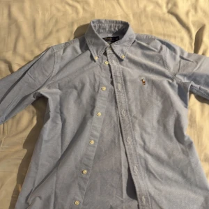 Ljusblå skjorta från Polo Ralph Lauren - Klassisk ljusblå skjorta från Polo Ralph Lauren i bomull. Skjortan har button-down krage, lång ärm och broderad logga på bröstet. Custom fit-modell med vita knappar och stilren look. Perfekt för dig som gillar tidlös och clean stil.