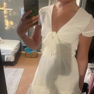 Studentklänning - Superfin vit kortklänning med djup v-ringning, knyt framtill och volangdetaljer. Klänningen har korta ärmar och en luftig passform, perfekt för varma dagar. Söt och trendig stil som passar till många tillfällen.