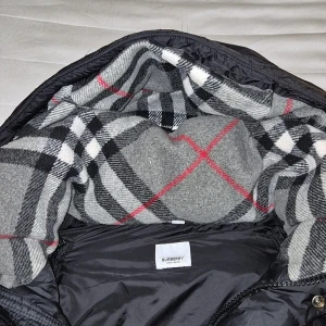 Svart pufferjacka från Burberry - Säljer en svart pufferjacka äkta från Burberry med klassiskt rutigt foder i grått, vitt, svart och rött. Jackan har dragkedja, knappar och två fickor framtill. Märkeslogga på bröstet och stor huva med rutigt innerfoder. Det kan även bli till en väst om man tar bort armarna genom dragkedjor inuti jackan