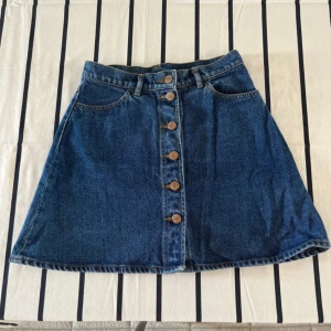 Jeanskjol med knappar från Monki - Säljer en klassisk blå jeanskjol från Monki i A-linjeformad modell. Kjolen har knappar framtill, två fickor bak och bälteshällor i midjan. Perfekt att styla med en enkel topp eller stickad tröja.