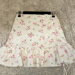 Blommig volangkjol från Gina Tricot - Supersöt kort kjol från Gina Tricot i vitt med rosa blommor och gröna blad. Kjolen har volangkant nertill och en lätt struktur i tyget. Perfekt för sommarens alla tillfällen och ger en romantisk vibe. Endast använd ett antal gånger och i stort sett som ny❤️💖💞