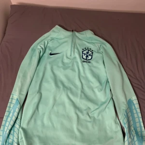 Nike Brasil långärmad fotbollströja M - Snygg ljusblå långärmad Brasilien-tröja från Nike med Dri-FIT-material. Tröjan har halv dragkedja, blått CBF-märke på bröstet och coolt rutmönster på ärmarna. Perfekt för dig som vill sticka ut på planen eller visa support för Brasilien.