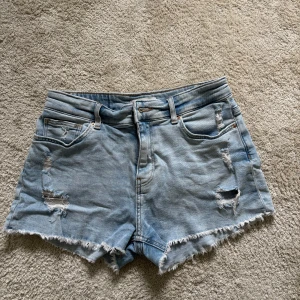 Ljusblå jeansshorts från H&M - Säljer ett par ljusblå jeansshorts från H&M med fransig kant och slitna detaljer framtill. Klassisk femficksmodell med normal passform och låg midja. Stretch material. 