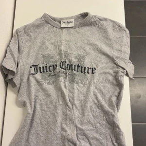 Grå t-shirt från Juicy Couture - Säljer en grå t-shirt från Juicy Couture med svart tryck framtill och diskret mönster i bakgrunden. Klassisk rund halsringning och korta ärmar. Perfekt för en avslappnad och trendig look.