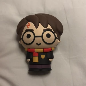 Powerbank Harry Potter med sladd - Rolig powerbank i form av Harry Potter med tillhörande blå laddkabel. Powerbanken har USB- och micro-USB-portar och är perfekt för att ladda mobilen på språng. Designen är inspirerad av Harry Potter med glasögon, blixtärr och Gryffindor-halsduk.