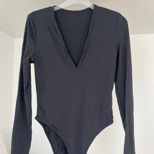 Svart långärmad bodysuit med djup v-ringning - Säljer en svart långärmad bodysuit med djup v-ringning. Perfekt att styla med jeans eller kjol för en trendig look. Mjuk och stretchig i materialet för en bekväm passform.