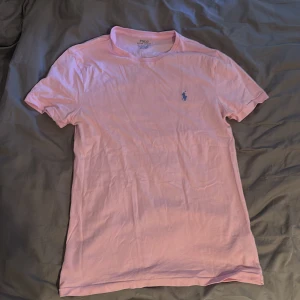 Rosa t-shirt från Polo Ralph Lauren - Snygg ljusrosa t-shirt från Polo Ralph Lauren med klassisk blå broderad logga på bröstet. Modellen är slim fit och har korta ärmar. Tillverkad i mjuk bomull som känns skön mot huden. Perfekt för en chill och stilren look.