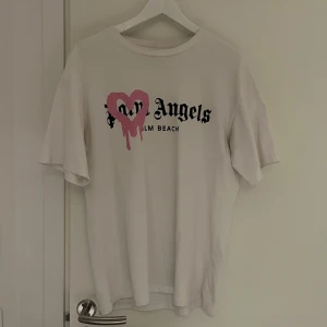 Vit Palm Angels t-shirt med tryck - Vit t-shirt från Palm Angels med svart text och ett rosa hjärta med drip-effekt på bröstet. Klassisk passform och rund halsringning. Perfekt statement-plagg för dig som gillar streetwear.