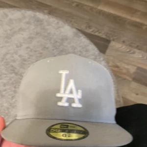 Grå LA-keps New Era 59FIFTY - Snygg grå keps från New Era 59FIFTY med LA-logga broderad framtill i vitt. Klassisk flat brim och MLB-logga baktill. Tillverkad i 100% polyester och passar dig som gillar streetwear och sportig stil.