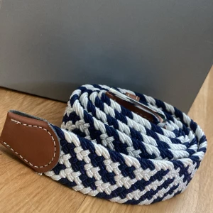 Randigt flätat bälte i blått och vitt - Snyggt flätat bälte med randigt mönster i marinblått och vitt. Bältet har detaljer i brunt skinn och ett silvrigt spänne. Perfekt accessoar för att lyfta din outfit med en sportig och fräsch känsla.
