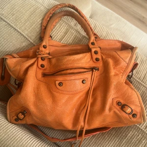 Orange handväska från Balenciaga - Cool orange handväska från Balenciaga med klassiska detaljer. Väskan är gjord i skinn och har flera fack samt dekorativa metalldetaljer. Perfekt för dig som vill sticka ut med en färgglad accessoar. NÅGRA DIFEKTER SOM JA TAGIT KORT PÅ! Har inget kvitto men den är äkta och tagit den till balenciaga o de kollade att den va älta, köpt på blocket.