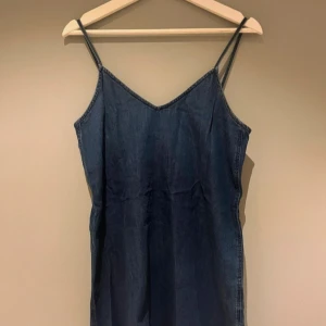 Jeansklänning med smala axelband - Snygg jeansklänning i mörkblå denim med smala axelband och v-ringning. Klänningen har metallringar bak som cool detalj och är ärmlös med en rak, något lös passform. Perfekt för en avslappnad och trendig look.