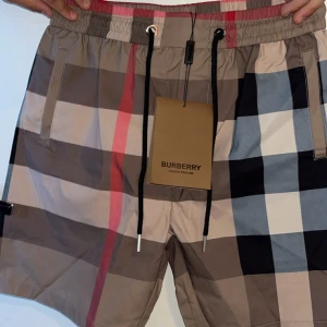 Burberry shorts  - Snygga shorts från Burberry med klassiskt rutigt mönster i beige, svart, vitt och rött. De har elastisk midja med svart snörning och praktiska fickor på sidorna. Perfekta för sommaren och ger en lyxig vibe till din outfit.