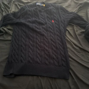 Svart kabelstickad tröja Polo Ralph Lauren - Kabelstickad svart tröja från Polo Ralph Lauren med klassisk röd broderad logga på bröstet. Tröjan har rund halsringning, långa ärmar och ribbade muddar. Perfekt för dig som gillar en tidlös och clean stil. Köpte den för 3 veckor sedan använd 3 gånger
