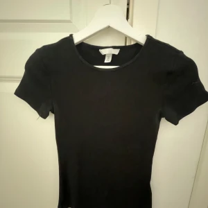 Svart ribbad topp från H&M - Enkel och stilren svart ribbad topp från H&M med rund halsringning och korta ärmar. Perfekt att matcha med jeans eller kjol för en clean look. Mjuk och stretchig för en bekväm passform.