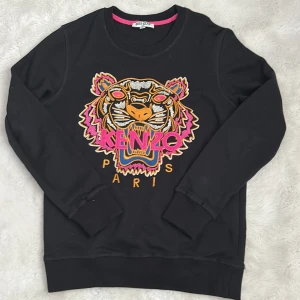 Svart Kenzo sweatshirt med tigerbrodyr - Svart sweatshirt från Kenzo med ikonisk broderad tiger och färgstarka detaljer i rosa, orange och blått. Tröjan är i 100% bomull, har rund hals och långa ärmar. Perfekt statement-plagg för dig som vill sticka ut.