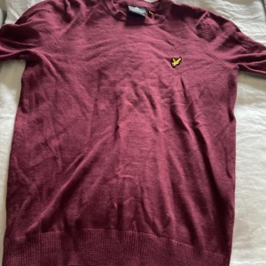 Vinröd stickad tröja från Lyle & Scott - Vinröd stickad tröja från Lyle & Scott med klassisk rund hals och broderad gul logga på bröstet. Tröjan har lång ärm och ribbade muddar. Perfekt för dig som gillar stilren och enkel design med en touch av färg.