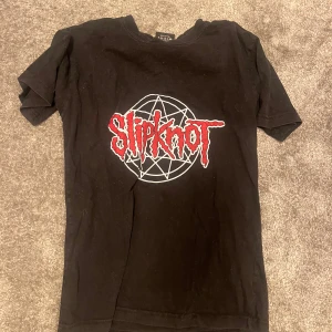 Svart Slipknot t-shirt med tryck - Svart t-shirt med stort Slipknot-tryck i rött och vitt framtill. Klassisk passform och rund hals. Perfekt för dig som gillar bandmerch och vill ha en edgy stil.