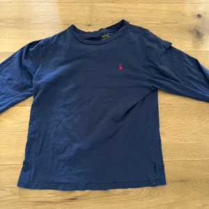 Blå långärmad tröja från Polo Ralph Lauren - Mörkblå långärmad tröja från Polo Ralph Lauren med rund halsringning och den klassiska röda logotypen broderad på bröstet. Perfekt för en stilren och avslappnad look. Passar kanske en S men troligen XS