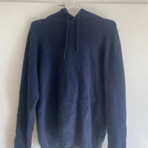 Mörkblå hoodie i kashmir från Soft Goat - Mörkblå hoodie från Soft Goat i kashmir. Tröjan har huva med snörning och långa ärmar.  Det är en herr model