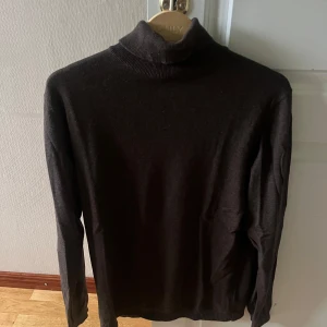 Mörkbrun polotröja i bomull - En stilren mörkbrun polotröja med lång ärm och klassisk hög krage. Tröjan är tillverkad i mjuk bomull och passar perfekt för lager-på-lager under kyliga dagar. Enkel att matcha med både jeans och kostymbyxor.