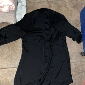 Svart morgonrock med spets H&M - Svart morgonrock från H&M i glansigt material med v-ringning. Klänningen har fina spetsdetaljer vid axlar och ärmslut samt trekvartsärm. Perfekt för dig som gillar en stilren look med lite extra detaljer.