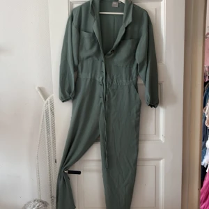 Grön jumpsuit med snörning - Stilren grön jumpsuit med långa ärmar, krage och knappar framtill. Dragsko i midjan för snygg passform och elastiska ärmslut. Perfekt för dig som gillar en avslappnad men ändå trendig look.