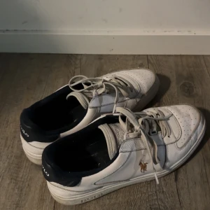 Vita sneakers från Polo Ralph Lauren - Snygga vita sneakers från Polo Ralph Lauren med klassisk logga på sidan. Skorna har marinblå detaljer vid hälen, perforerad tå och snörning framtill. Tillverkade i skinn och syntet för en sportig och stilren look. OBS!! SKORNA KOMMER SÅKLART FINA OCH TVÄTTADE !