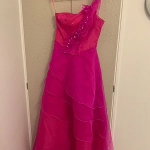 Rosa långklänning med volang och pärlor - Snygg rosa långklänning i glansigt material med ett axelband och volangdetaljer längs ena axeln. Klänningen har pärlor och paljetter på överdelen samt lager av transparent tyg som ger en cool effekt. Perfekt för bal eller fest och har dragkedja i ryggen. Köpt i Tyskland. Använt 1 gång. 