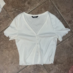 Vit ribbad topp med knappar från SHEIN - Säljer en söt vit ribbad topp från SHEIN i storlek XS. Toppen har korta ärmar, v-ringning och tre knappar framtill. Den är croppad med vågig kant nertill och vid ärmslut, vilket ger en trendig look. Perfekt att styla med jeans eller kjol.