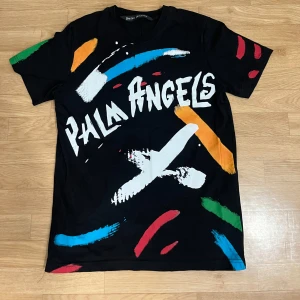 Palm Angels svart t-shirt med färgstänk - Svart t-shirt från Palm Angels med stora vita loggan framtill och coola färgstänk i blått, rött, grönt, orange och vitt. Kortärmad modell i mjuk bomull, perfekt för dig som gillar streetwear med attityd.