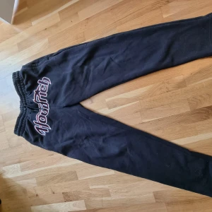 Svarta mjukisbyxor från Hoodrich - Säljer ett par svarta mjukisbyxor från Hoodrich med stor broderad logga i rött och vitt över framdelen. Byxorna har resår och snörning i midjan för skön passform. Perfekta för chill eller streetwear-stil.